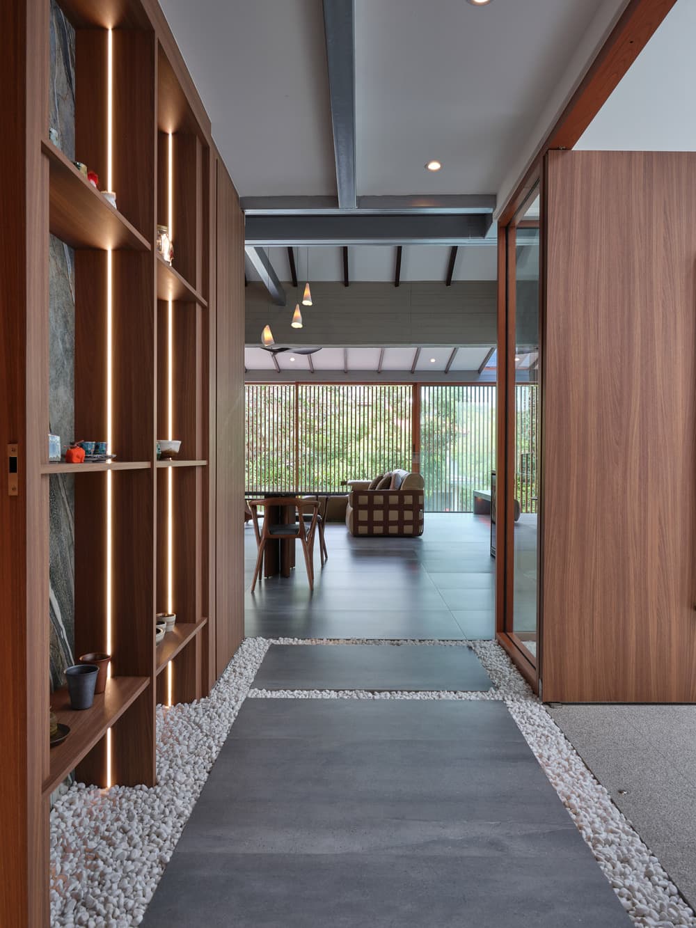 The stone path between beds of white pebbles, past a lit timber shelf of ceramics to the dining room, at A Sensory House, a luxury landed house interior in Singapore by K2SD Group